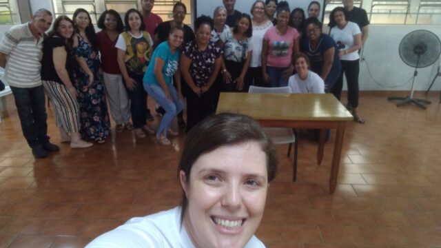 Descrição da imagem: foto do tipo selfie com 8 pessoas, sendo dois homens e seis mulheres, todos em pé com um fundo cinza e algumas cadeiras pretas entre as pessoas.Fim da descrição