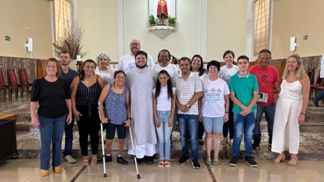 #paratodosverem Foto de um grupo de 16 pessoas, entre homens e mulheres de diferentes idades. Dentre elas há um sacerdote com vestes litúrgicas roxa e algumas pessoas com deficiência. Elas estão organizadas em duas fileiras, uma ao lado da outra em frente ao presbitério de uma Igreja com paredes e piso em tons claro, nas laterais há algumas cadeiras de madeira com estofado vermelho, também há um ar condicionado e uma janela em cada parede lateral. Na parede dos fundos há um altar com a imagem de Nosso Senhor Bom Jesus. Fim da descrição.