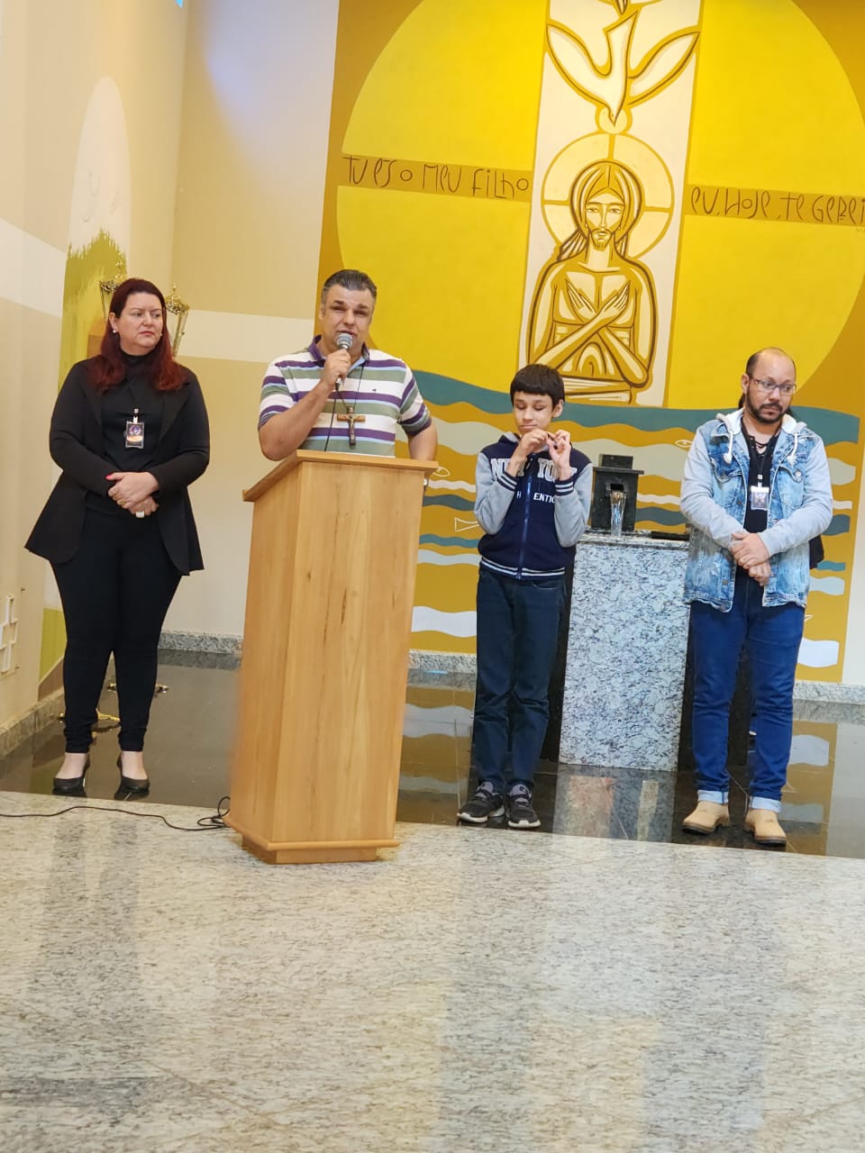 Descrição da Imagem: Foto de um grupo de quatro pessoas, uma ao lado da outra próximas a uma pia batismal. Da esquerda para a direita há uma mulher, pele clara, cabelos ruivo na altura do ombro, veste roupa e sapato preto. Um homem com camisa com listras na horizontal nas cores branca, azul e verde, ele está atrás de um ambão e segura um microfone com a mão direita. Um menino, pele clara, cabelo escuro, usa agasalho azul marinho e cinza, calça jeans e tênis escuro. Um homem, pele clara, um pouco careca, usa óculos, camisa preta, casaco e calça jeans e sapato marrom. Observa-se um piso de granito e ao fundo um painel em tons de amarelo com a cena do batismo do Senhor. Fim da Descrição.