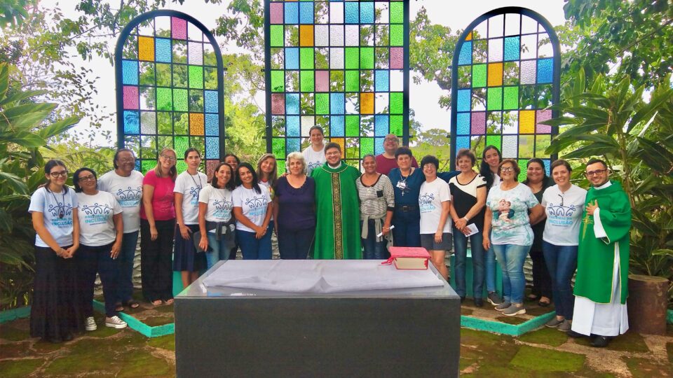 Descrição da Imagem: Foto de um grupo de 22 pessoas de várias idades, sendo 19 mulheres e 3 homens. Algumas mulheres usam uma camisa branca com a logo da Pastoral da Inclusão e 2 homens usam veste litúrgica na cor verde. Eles estão um ao lado do outro, sorrindo para a foto. Na frente deles há um altar de mármore escuro com uma toalha branca e um livro litúrgico de capa vermelha. Atrás do grupo há 3 painéis com vitrais retangulares nas cores azul, verde, amarelo e rosa. Ao fundo observa-se algumas árvores e o céu. Fim da descrição.