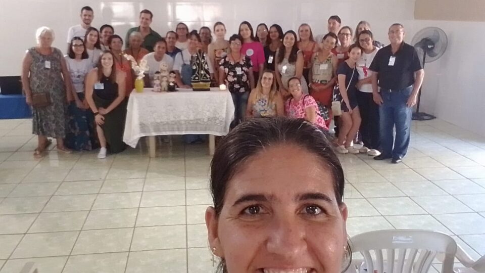 Descrição da imagem: Fotografia estilo selfies de uma mulher em primeiro plano com um grupo de aproximadamente 30 pessoas. A mulher em primeiro plano está sorrindo, possui cabelos castanho preso para trás. Ao fundo o grupo de pessoas é composto por homens e mulheres de diversas faixas etárias. Estes estão atrás de uma mesa com uma toalha de renda branca, acima da mesa há uma vela, uma imagem de nossa senhora aparecida e um vaso de orquídea branca. Fim da descrição.